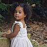 toddler, child, curly_hair, white_dress, outdoor, stone_wall, greenery, leaves, nature, portrait, young_girl, standing, serene, cute, fashion, summer, innocence, person, garden, soft_light