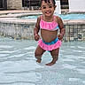 toddler, child, smiling, swimsuit, pool, water, curly_hair, outdoor, summer, backyard, happy, standing, playful, shallow_water, water_droplets, stone_wall, chair, grill, sunny, cute