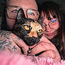 cat, tortoiseshell_cat, pet, yellow_eyes, glasses, human_faces, couple, tattoo, hand, collar, bell, whiskers, close_up, indoor, soft_blanket, smile, brown_hair, facial_hair, portrait, cozy