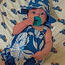 baby, pacifier, blue_dress, hat, bed, sheet, pattern, trucks, infant, clothing, lying_down, cute, wide_eyes, hand, foot, indoors, child, face, sleepwear, comfort