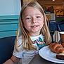 blonde_hair, bracelet, breakfast, cafe, cake, chair, child, croissant, dining, food, furniture, happy, indoor, light, person, plate, portrait, smile, table, tshirt