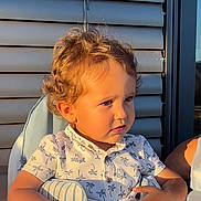 Lyzio a rejoint le concours — aidez-le/la à gagner de superbes lots ! child, toddler, curly_hair, blond_hair, portrait, sunlight, high_chair, seat, patterned_shirt, palm_tree_shirt, closeup, outdoor, balcony, window_shutters, face, pensive_expression, summer, skin, arm, casual_clothing