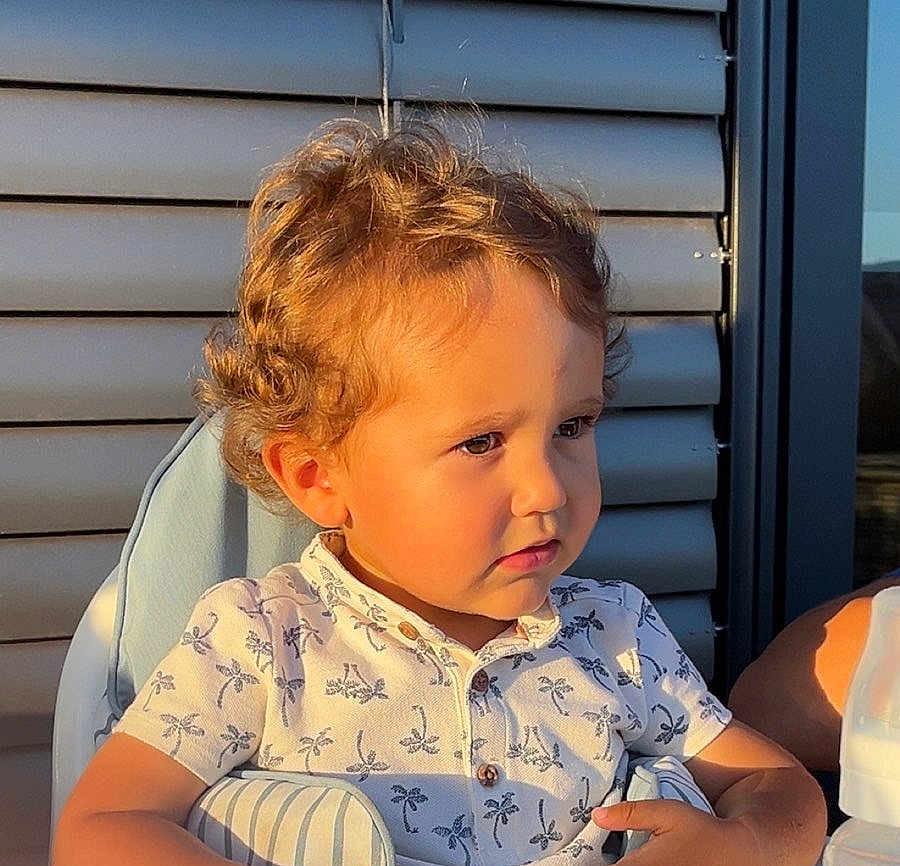Lyzio a rejoint le concours — aidez-le/la à gagner de superbes lots ! child, toddler, curly_hair, blond_hair, portrait, sunlight, high_chair, seat, patterned_shirt, palm_tree_shirt, closeup, outdoor, balcony, window_shutters, face, pensive_expression, summer, skin, arm, casual_clothing