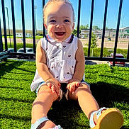 Amani is registered to the contest to win money with this photo: child, toddler, smiling, sitting, outdoor, sunlight, grass, balcony, curly_hair, white_top, sandals, happy, face, legs, fence, daylight, nature, person, summer, casual