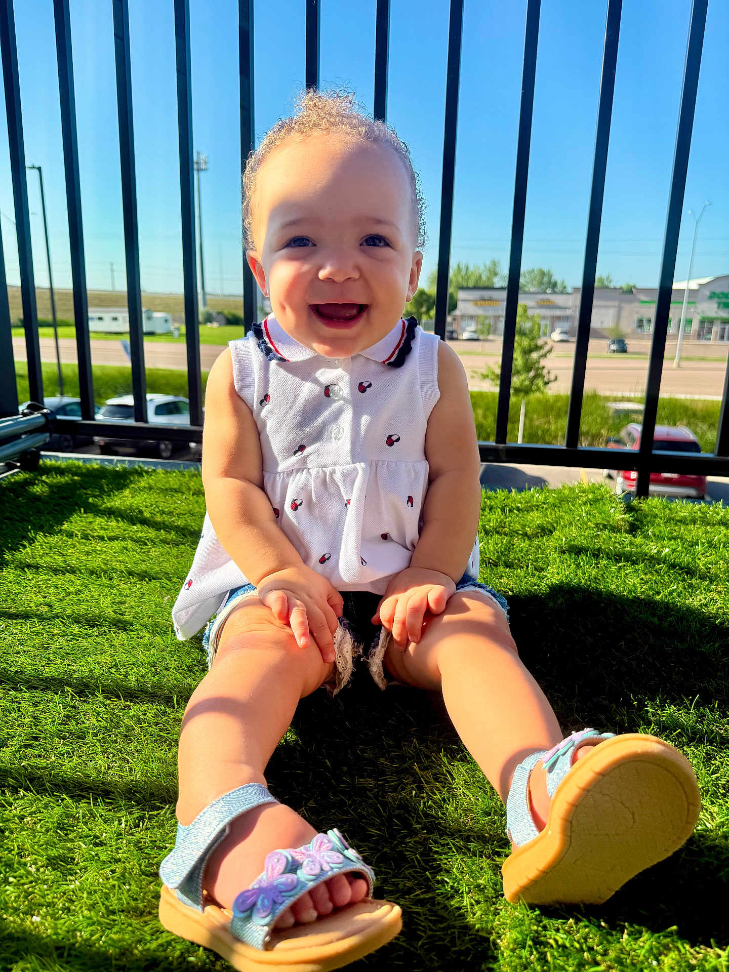 Amani is registered to the contest to win money with this photo: child, toddler, smiling, sitting, outdoor, sunlight, grass, balcony, curly_hair, white_top, sandals, happy, face, legs, fence, daylight, nature, person, summer, casual
