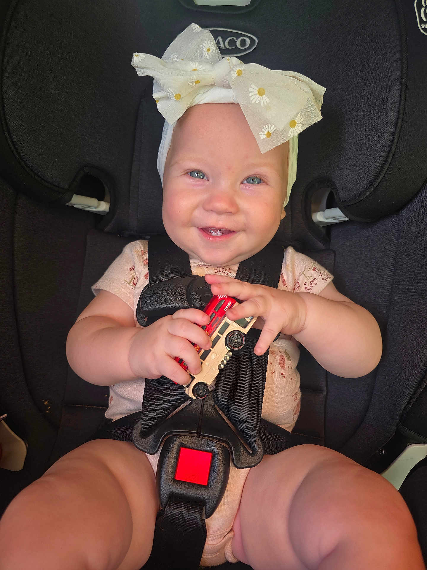 Alruna is registered to the contest to win money with this photo: baby, child, car_seat, toy, bus_toy, smiling, headband, bow, happy, infant, safety_harness, cute, chubby_cheeks, clothing, seatbelt, indoor, person, closeup, hands, face