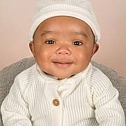 Saint is registered to the contest to win money with this photo: baby, infant, smile, white_sweater, knit_hat, cute, portrait, child, face, happy, warm_clothing, indoor, soft_lighting, cozy, young_child, close_up, headwear, buttoned_sweater, neutral_background, adorable