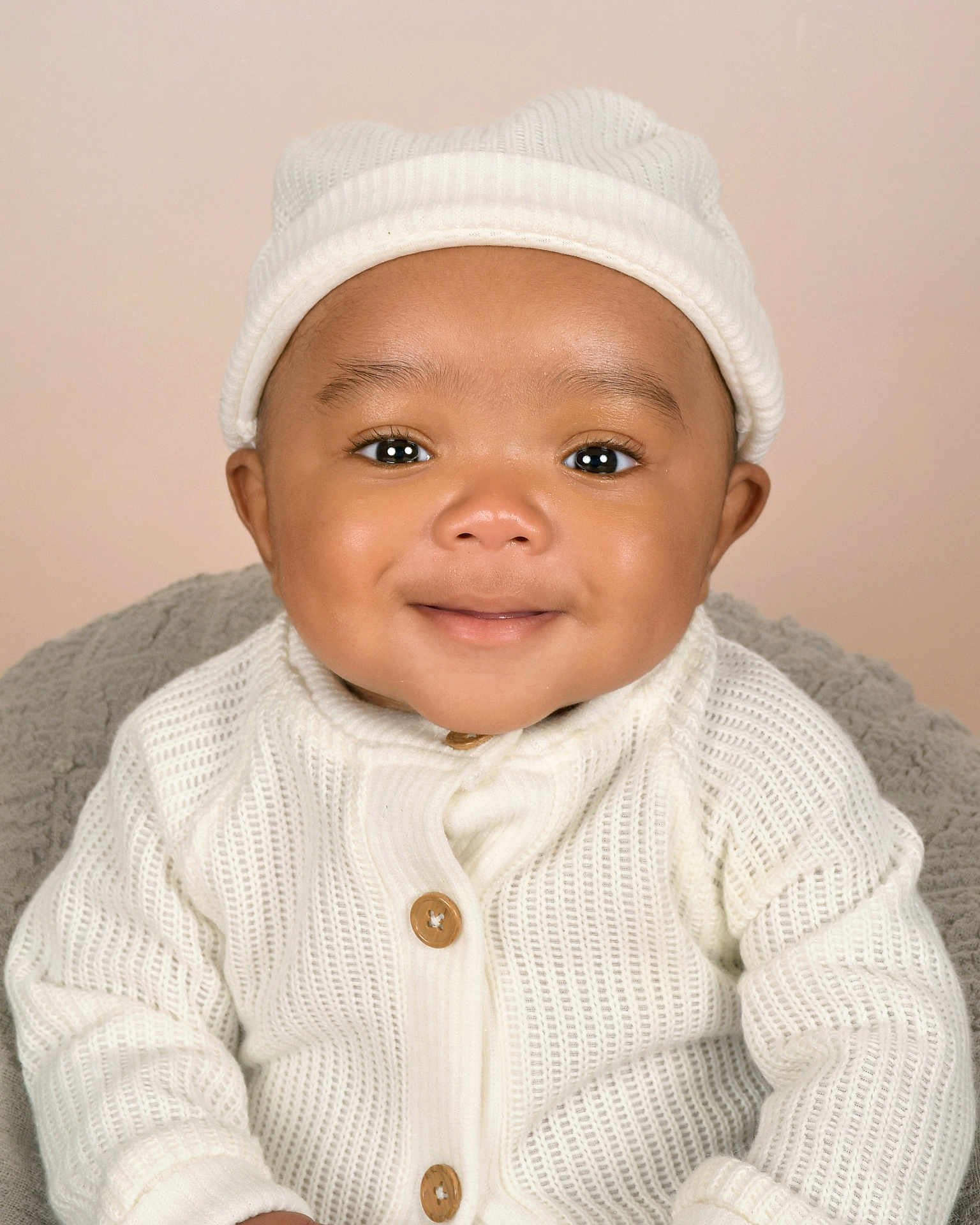 Saint is registered to the contest to win money with this photo: baby, infant, smile, white_sweater, knit_hat, cute, portrait, child, face, happy, warm_clothing, indoor, soft_lighting, cozy, young_child, close_up, headwear, buttoned_sweater, neutral_background, adorable