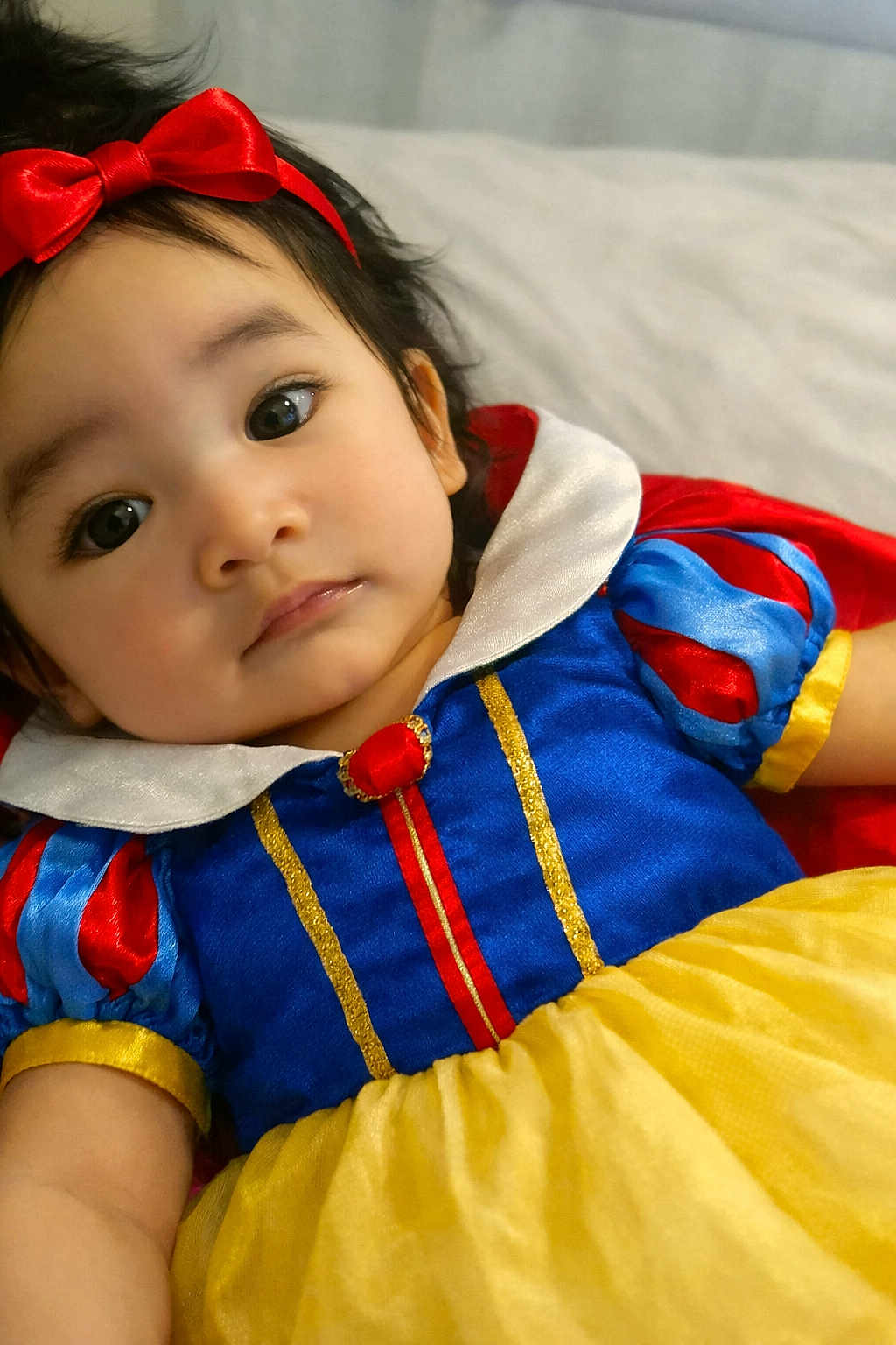 Sara joined the competition — help win amazing prizes! baby, infant, child, face, eyes, headband, bow, costume, snow_white_costume, dress, collar, colorful, fabric, portrait, lying_down, blanket, close_up, cute, gaze, cheeks