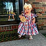 Mckenzie is registered to the contest to win money with this photo: baby, child, girl, dress, headband, bow, sandals, porch, brick_wall, welcome_mat, wooden_sign, outdoor, smiling, happy, person, standing, summer, reflection, window, concrete