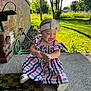 Mckenzie is registered to the contest to win money with this photo: toddler, child, dress, headband, bow, plaid, outdoor, porch, concrete, brick_wall, greenery, sunlight, grass, trees, happy, smiling, holding, token, shoe, playful