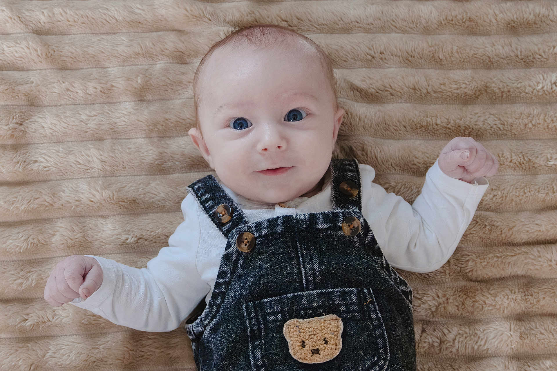 Naïm participe au concours pour gagner de l'argent avec cette photo : baby, infant, denim_overalls, teddy_bear_patch, blue_eyes, cute, smile, lying_down, soft_blanket, beige, clothing, child, portrait, closeup, happy, newborn, skin, face, person, indoors