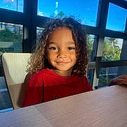 Nathanael participe au concours pour gagner de l'argent avec cette photo : child, curly_hair, smile, red_shirt, indoor, table, window, sunlight, blue_sky, greenery, portrait, face, person, happy, young_child, natural_light, seated, casual, daylight, interior
