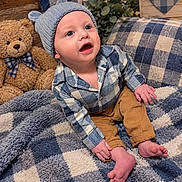 Corey is registered to the contest to win money with this photo: baby, child, infant, portrait, smiling, wide_eyed, indoor, seated, plaid, hat, shirt, pants, bare_feet, teddy_bear, blanket, couch, rustic_decor, plant, heart_decoration, cozy
