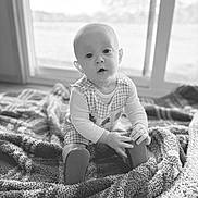 Hudson is registered to the contest to win money with this photo: baby, child, infant, blanket, indoor, window, holding_object, sitting, curious, monochrome, soft_light, face, hands, footwear, long_sleeve, checkered_clothing, floor, home, portrait, young