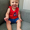 Harper is registered to the contest to win money with this photo: baby, child, smiling, sitting, red_shirt, blue_shorts, couch, indoor, happy, cute, infant, person, furniture, foot, hand, face, skin, expression, young, casual
