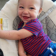 Harper is registered to the contest to win money with this photo: baby, child, smile, happy, face, striped_shirt, blue_shorts, cushion, baby_seat, indoor, person, young_child, cute, infant, toddler, clothing, seatbelt, skin, arm, hair