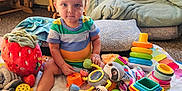 Logan is registered to the contest to win money with this photo: baby, blanket, blue_eyes, carpet, child, colorful, cushion, cute, floor, indoor, playtime, plush, seat, soft_toys, stacking_rings, steering_wheel_toy, striped_shirt, stuffed_animal, toddler, toys