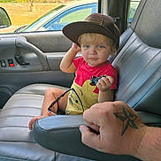 Logan is registered to the contest to win money with this photo: child, toddler, toy_car, baseball_cap, car_interior, car_seat, tattoo, adult_hand, window, greenery, parked_car, red_shirt, yellow_shirt, sunglasses, foot, armrest, door_panel, seat_belt, person, portrait