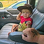 child, toddler, toy_car, baseball_cap, car_interior, car_seat, tattoo, adult_hand, window, greenery, parked_car, red_shirt, yellow_shirt, sunglasses, foot, armrest, door_panel, seat_belt, person, portrait
