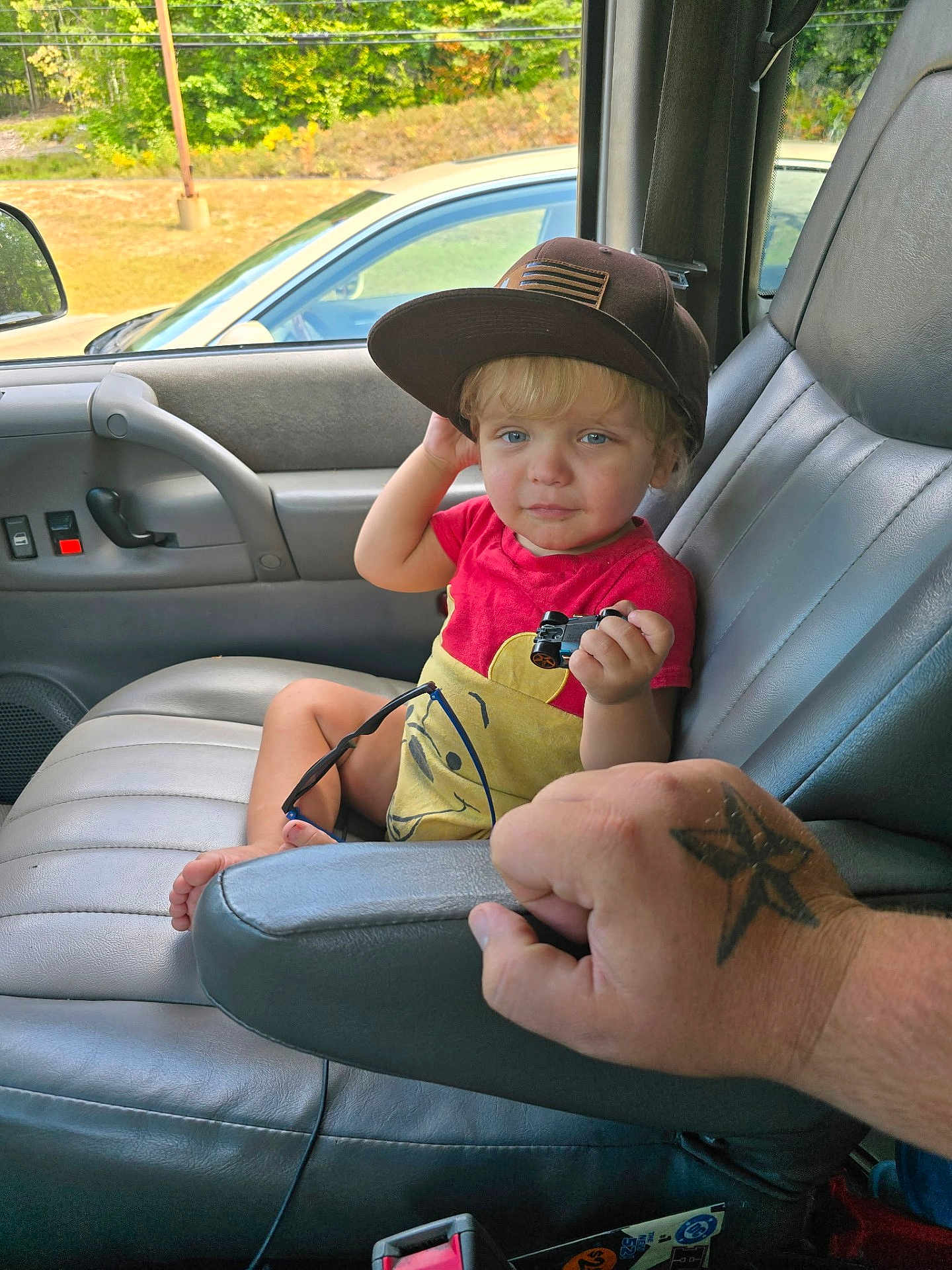 Logan is registered to the contest to win money with this photo: child, toddler, toy_car, baseball_cap, car_interior, car_seat, tattoo, adult_hand, window, greenery, parked_car, red_shirt, yellow_shirt, sunglasses, foot, armrest, door_panel, seat_belt, person, portrait