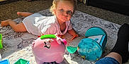 Logan is registered to the contest to win money with this photo: blocks, blue_eyes, books, carpet, child, floor, furniture, indoor, leg, person, piggy_bank, plastic_toys, room, shorts, sock, stuffed_animals, sunlight, toddler, toy, white_clothing