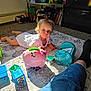 Logan is registered to the contest to win money with this photo: toddler, child, toy, piggy_bank, blocks, carpet, sunlight, indoor, blue_eyes, white_clothing, person, furniture, books, stuffed_animals, plastic_toys, floor, leg, shorts, sock, room