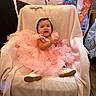 baby, child, pink_dress, chair, blanket, party, celebration, smile, happy, indoor, footwear, person, portrait, cute, toddler, decorations, wooden_floor, jewelry, ribbon, fabric