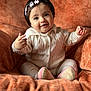armchair, baby, child, cozy, cute, footwear, hair_clips, happy, indoor, infant, knitted_clothing, orange, person, plush, portrait, seated, small_hands, smiling, soft_lighting, warm_colors