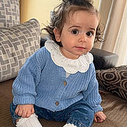 Mathilde a rejoint le concours — aidez-le/la à gagner de superbes lots ! toddler, child, indoor, couch, blue_cardigan, white_socks, curly_hair, plant, pillow, cozy, casual_clothing, cute, portrait, sitting, home, soft_lighting, curious_expression, baby, denim_pants, earrings