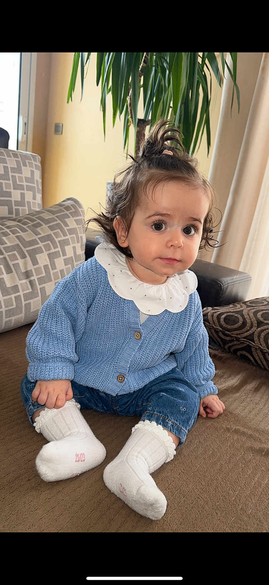 Mathilde a rejoint le concours — aidez-le/la à gagner de superbes lots ! toddler, child, indoor, couch, blue_cardigan, white_socks, curly_hair, plant, pillow, cozy, casual_clothing, cute, portrait, sitting, home, soft_lighting, curious_expression, baby, denim_pants, earrings