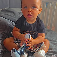 Kylah joined the competition — help win amazing prizes! toddler, child, bed, blanket, controller, toy, socks, clothing, indoor, furniture, crib, person, face, hair, expression, skin, hand, footwear, sitting, curious