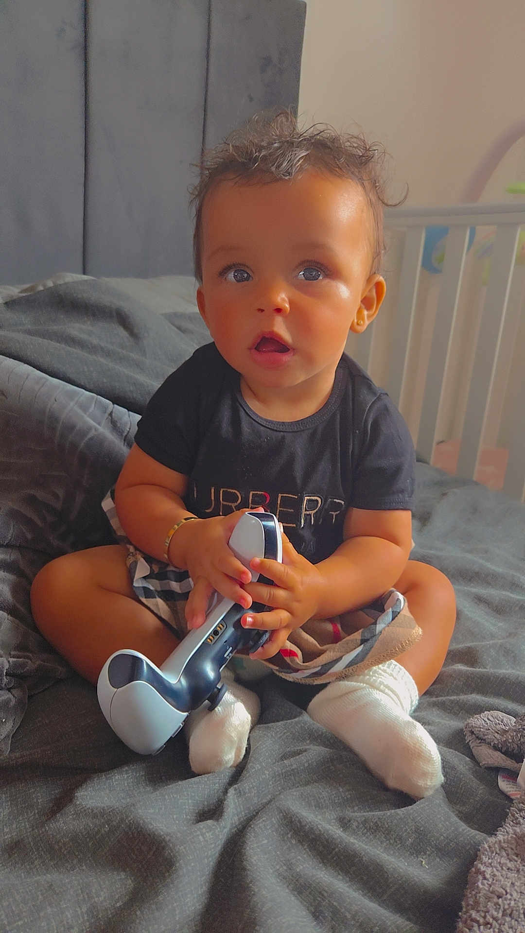 Kylah joined the competition — help win amazing prizes! toddler, child, bed, blanket, controller, toy, socks, clothing, indoor, furniture, crib, person, face, hair, expression, skin, hand, footwear, sitting, curious