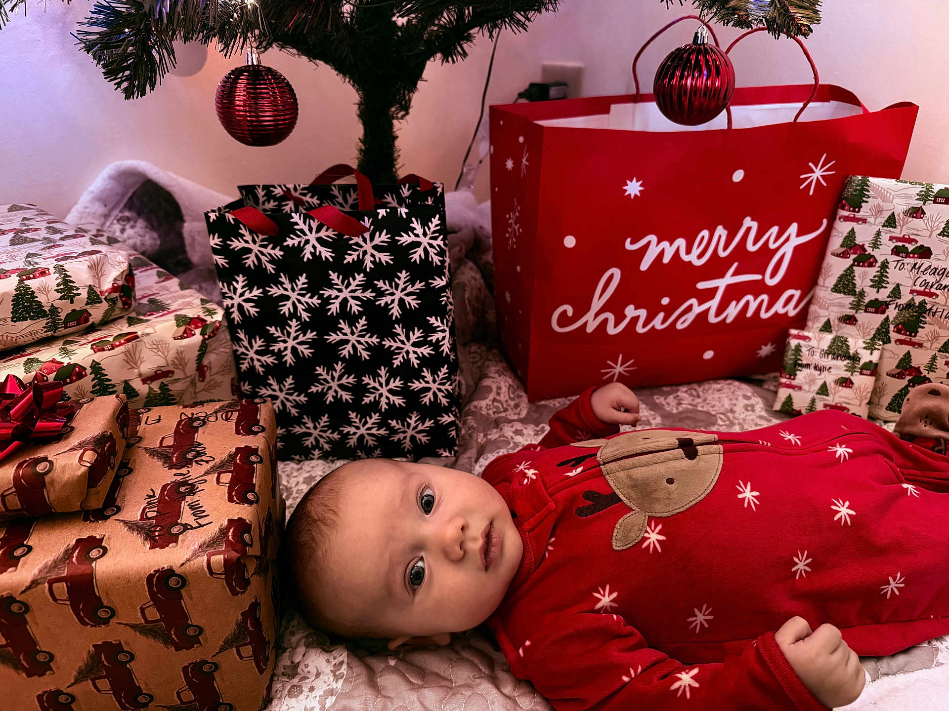 Atlas joined the competition — help win amazing prizes! baby, christmas, presents, gift_bags, red_onesie, reindeer_design, holiday, festive, decorated_tree, ornaments, wrapped_gifts, indoor, cozy, infant, lying_down, curious, holiday_decor, celebration, winter, festivity