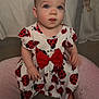 Imogen is registered to the contest to win money with this photo: baby, child, dress, ladybug_pattern, red_bow, pink_cushion, playpen, curious, sitting, indoor, soft_furniture, face, person, infant, cute, clothing, head, expression, comfort, looking_up