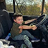 child, boy, steering_wheel, car_interior, seat, sneakers, window, sunlight, smartphone, paper_towel, casual_clothing, playful, vehicle, seatbelt, dashboard, seat_cover, grip, daylight, smile, indoor