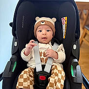 Arlo is registered to the contest to win money with this photo: baby, child, car_seat, hat, bear, checkered_pants, socks, straps, sitting, indoors, wooden_chair, floor, cute, curious, infant, clothing, person, face, hands, safety