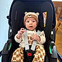 baby, child, car_seat, hat, bear, checkered_pants, socks, straps, sitting, indoors, wooden_chair, floor, cute, curious, infant, clothing, person, face, hands, safety
