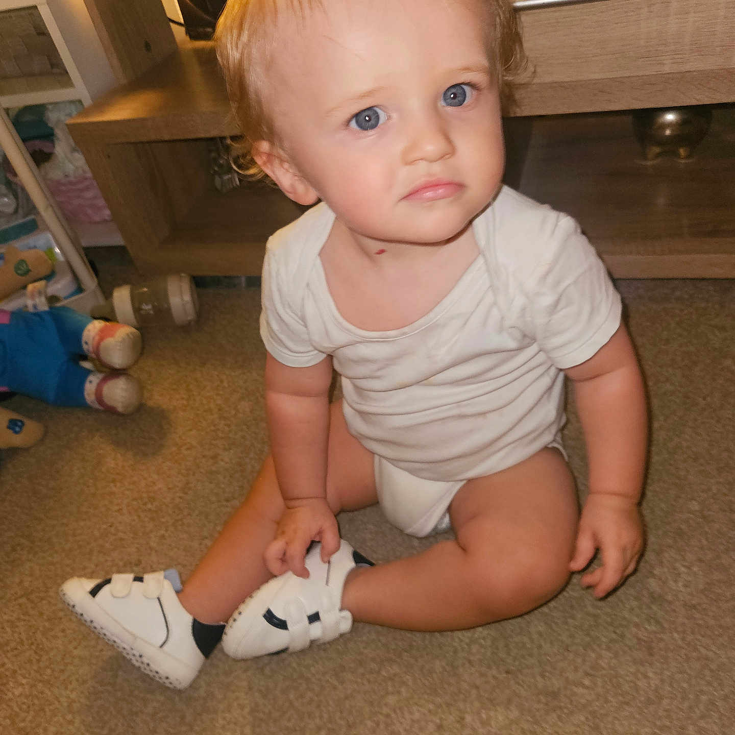 Anthony-Junior joined the competition — help win amazing prizes! blonde_hair, blue_eyes, carpet, child, curly_hair, cute, expression, floor, furniture, home, indoor, infant, person, sitting, toddler, toy, white_onesie, white_sneakers, wooden_shelf, young_child