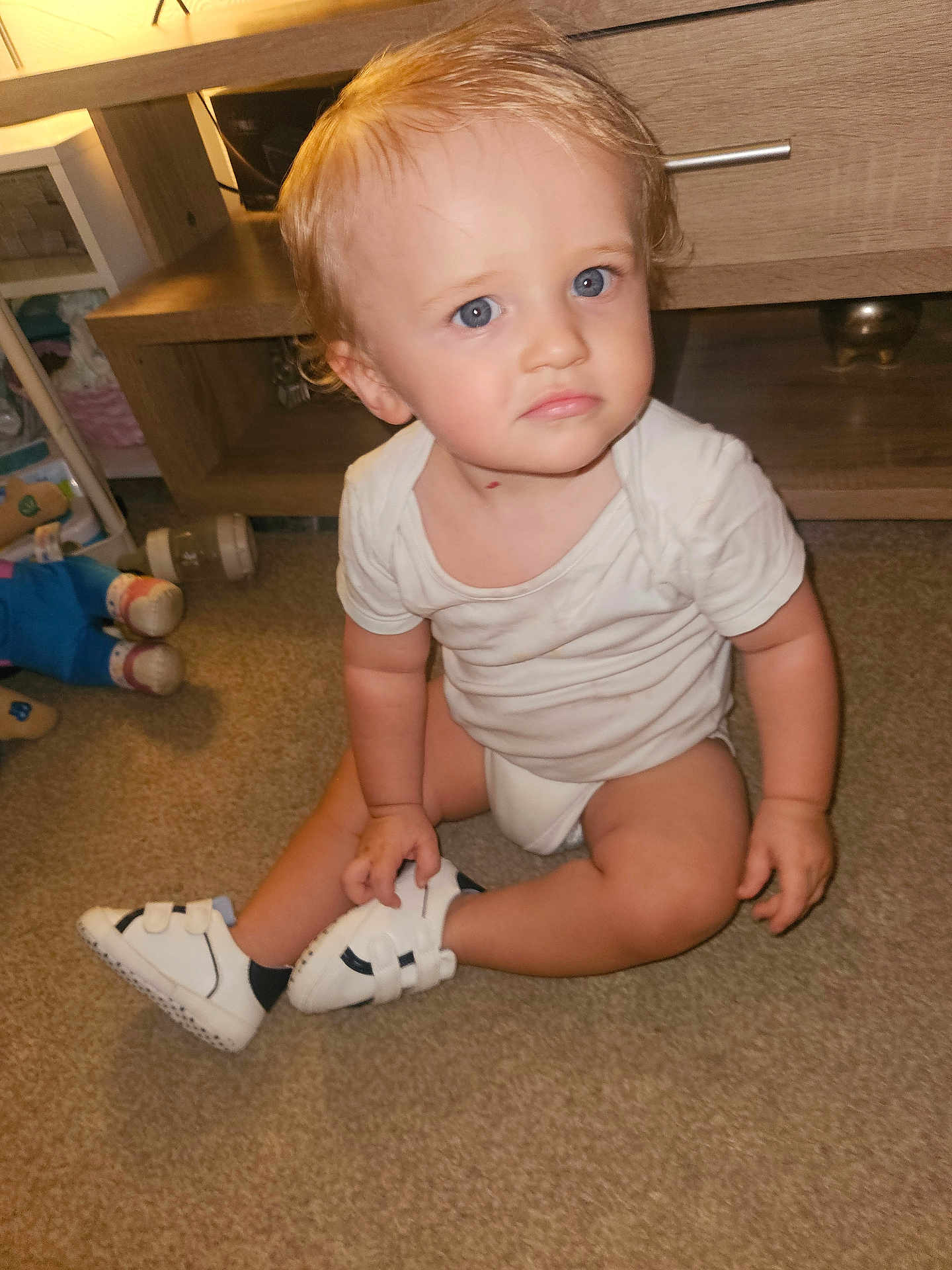Anthony-Junior joined the competition — help win amazing prizes! blonde_hair, blue_eyes, carpet, child, curly_hair, cute, expression, floor, furniture, home, indoor, infant, person, sitting, toddler, toy, white_onesie, white_sneakers, wooden_shelf, young_child