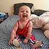 Oakley joined the competition — help win amazing prizes! baby, child, smiling, red_clothing, blue_clothing, white_clothing, bed, bedspread, indoor, happy, person, portrait, sitting, footwear, plaid_pattern, furniture, blanket, skin, head, cute