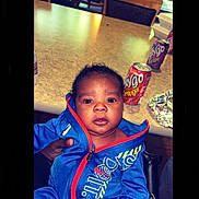 Jarvis is registered to the contest to win money with this photo: baby, child, blue_jacket, sitting, table, soda_cans, orange_soda, grape_soda, indoor, curly_hair, face, eyes, hand, clothing, person, portrait, expression, curious, food, drink