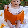 Harper is registered to the contest to win money with this photo: baby, pumpkin, candy_corn, outdoor, grass, autumn, child, white_clothing, sitting, expression, cute, festive, orange, leaf_litter, hand, foot, person, skin, head, nature