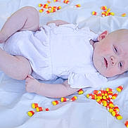 Harper is registered to the contest to win money with this photo: baby, infant, candy_corn, white_outfit, lying_down, soft_lighting, pale_skin, blue_eyes, chubby_arms, chubby_legs, sheet, background, indoors, cute, child, portrait, food, colorful, sweet, holiday