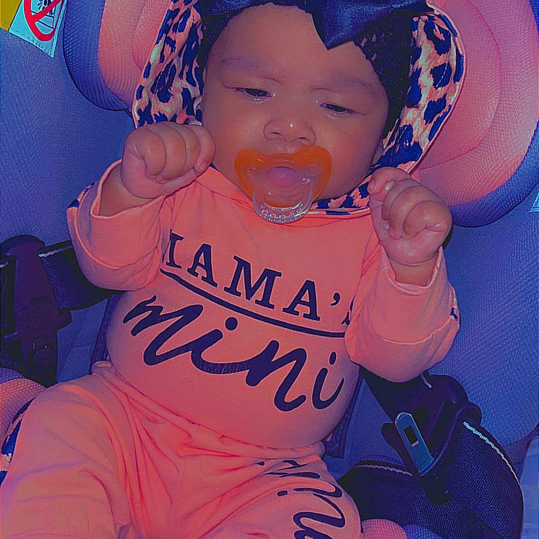 Elianna is registered to the contest to win money with this photo: baby, baby_girl, bow, car_seat, child, clothing, cute, headwear, indoors, infant, orange_outfit, pacifier, person, portrait, safety, seatbelt, sitting, socks, text_on_clothing, young_child