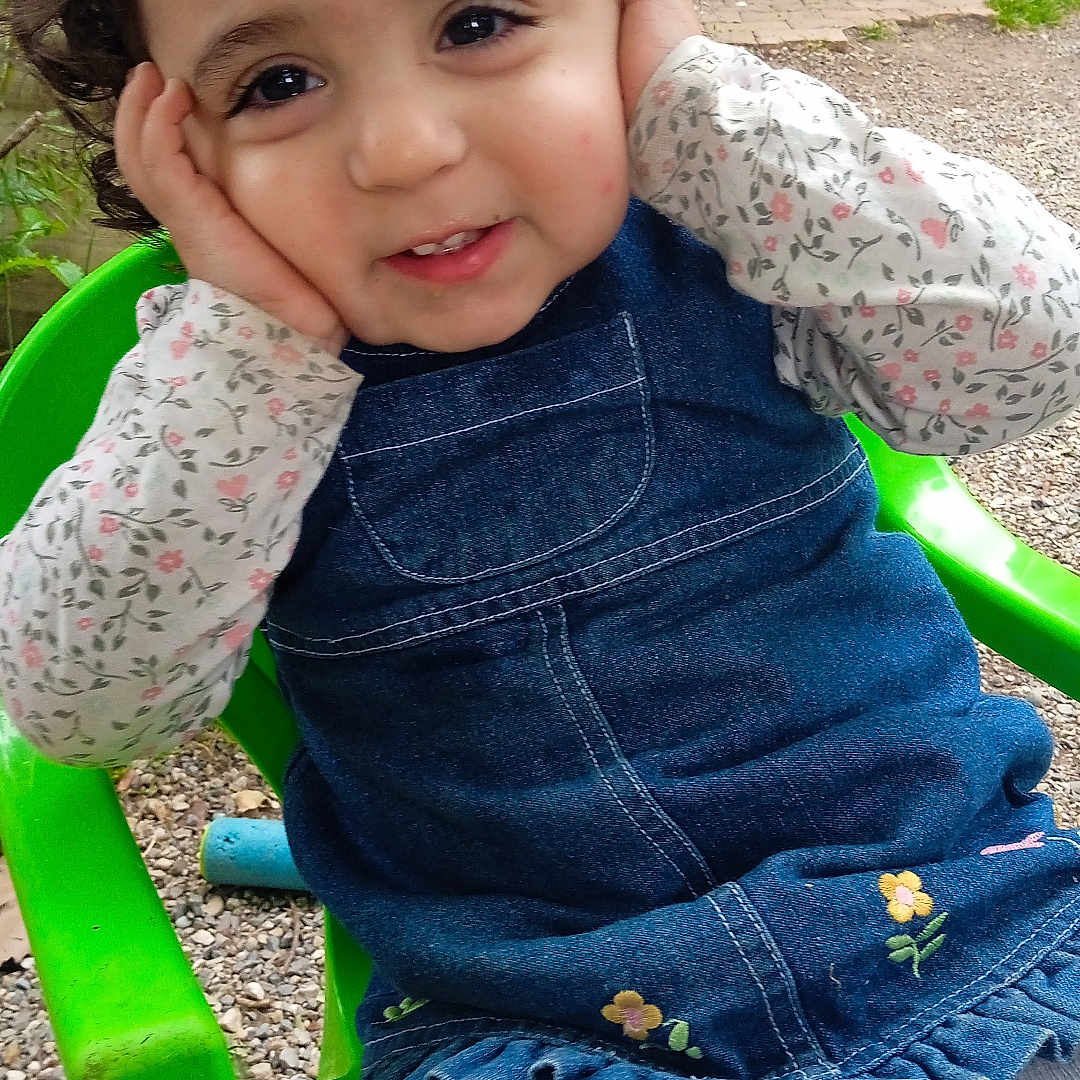 Julia participe au concours pour gagner de l'argent avec cette photo : casual_clothing, child, curly_hair, daylight, denim_dress, face, floral_pattern, garden, green_chair, ground, hands, happy, nature, outdoor, person, playful, seated, smiling, toddler, white_tights