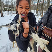 Vera is registered to the contest to win money with this photo: animal, baby_goat, blue_teeth, casual_clothing, child, daylight, farm, fun, goat, happy, holding, jacket, nature, outdoor, person, playful, smiling, snow, trees, winter