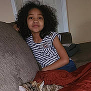 Vera is registered to the contest to win money with this photo: child, girl, curly_hair, striped_shirt, couch, sofa, living_room, cat, sleeping_cat, blanket, red_blanket, pillow, portrait, indoor, sitting, relaxed, door, dark_hair, pet, smile