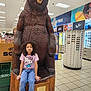 child, girl, bear_statue, wood_carving, retail_store, sunglasses_display, shopping, portrait, indoor, tile_floor, hello_kitty, jeans, casual_clothing, store_signage, ceiling_lights, shopper, display_rack, posing, statue, store_interior