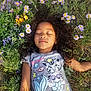 child, girl, sleeping, flowers, wildflowers, meadow, grass, curly_hair, t_shirt, mermaid_graphic, cat_graphic, necklace, closed_eyes, portrait, outdoors, nature, relaxed, lying_down, sunlight, colorful